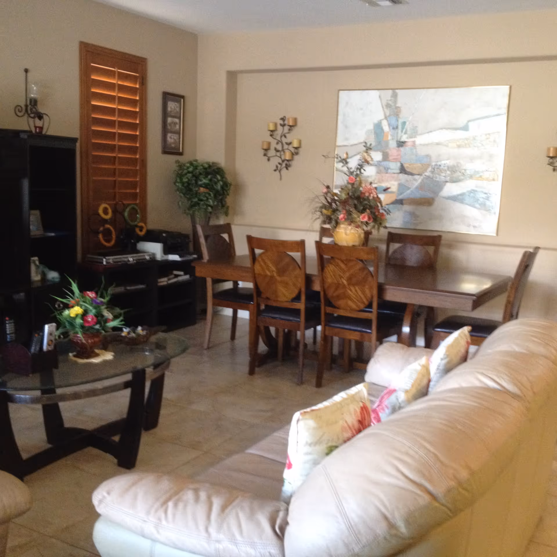 Interior view of a living and dining area featuring a beige leather sofa with floral cushions in the foreground, a glass coffee table with a flower arrangement, a wooden dining table with six chairs, decorative wall art, and a potted plant near a window with wooden shutters.