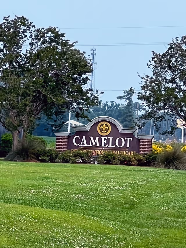 A landscaped outdoor area with a large sign reading 'CAMELOT' in white letters on a dark background, flanked by two trees and surrounded by green grass and bushes.