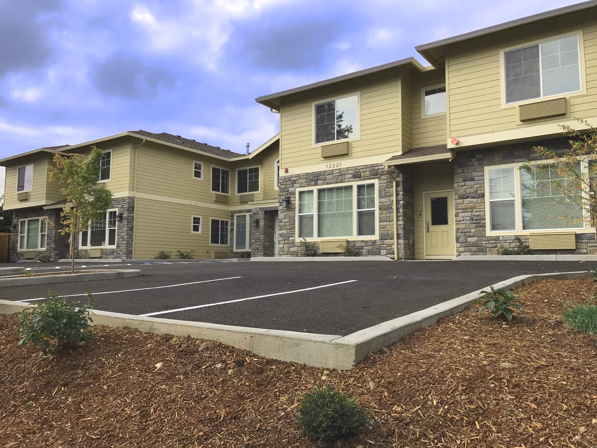 Exterior view of a two-story residential building with beige siding and stone accents, surrounded by a parking lot and landscaped areas with small trees and shrubs under a cloudy sky.