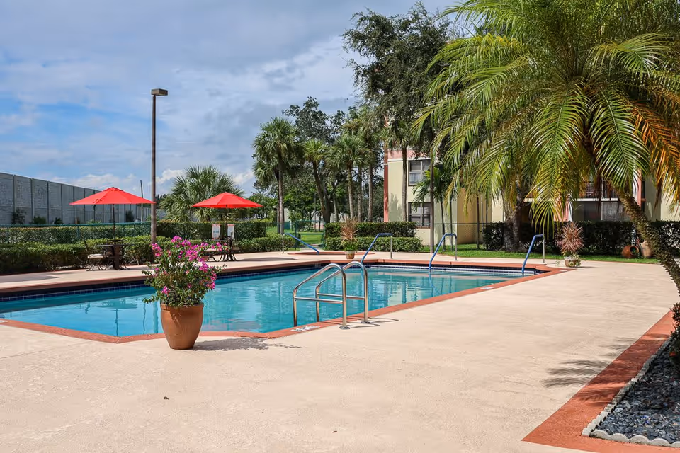 Outdoor swimming pool area with clear blue water, surrounded by a beige concrete deck. There are two red umbrellas shading tables and chairs on the left side, potted plants with pink flowers near the pool, palm trees, and a building in the background under a partly cloudy sky.