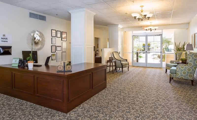 Reception area of a senior living facility with a wooden front desk on the left, a computer and a potted orchid on the desk, framed certificates on the wall behind, and a seating area with patterned armchairs and a small table near glass double doors leading outside.