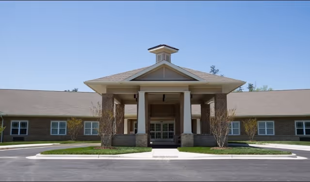 Front exterior view of Chatham Ridge Assisted Living facility showing a single-story brick building with a covered entrance supported by four columns, surrounded by small landscaped areas and a paved driveway under a clear blue sky.