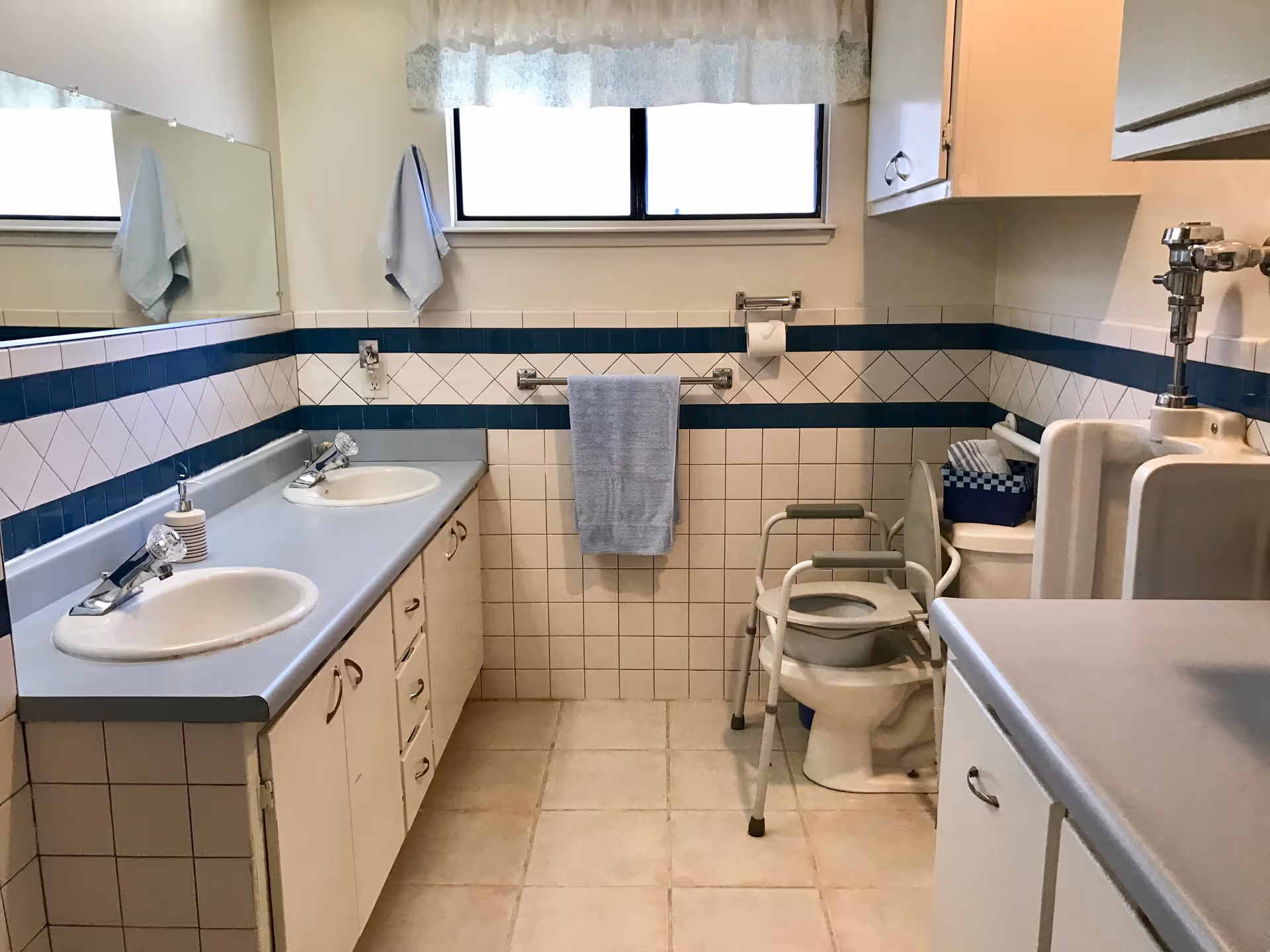 A clean bathroom with two sinks on the left side, a large mirror above the sinks, a window with a lace curtain, a towel rack with a blue towel, a toilet with a raised seat and support frame, a urinal, and cabinets on the right side. The walls are tiled with white and blue tiles.