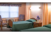 Two twin beds with green bedspreads in a softly lit room featuring a nightstand with flowers, two chairs by a window, and wall-mounted lighting.