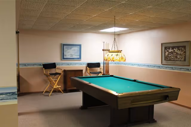 A recreational room with a pool table in the center, two director-style chairs against the wall, a small cabinet between the chairs, a hanging stained glass lamp above the pool table, and framed artwork on the walls.
