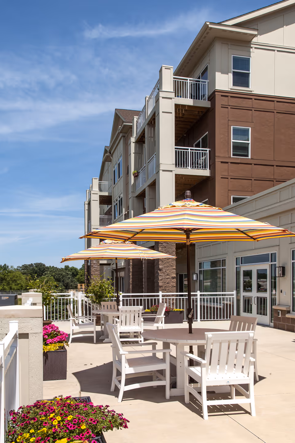 Outdoor patio area at The Waters on Mayowood with white tables and chairs under large striped umbrellas, surrounded by flower planters and adjacent to a multi-story building with balconies.