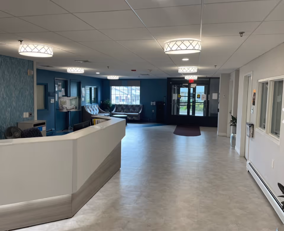 Interior view of a senior living facility lobby with a white reception desk on the left, blue accent walls, modern ceiling lights, seating area with couches near a window, and glass entrance doors at the far end.