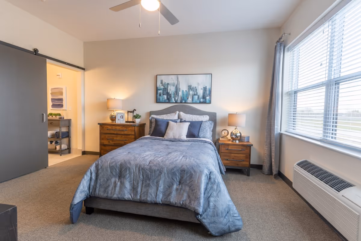 A neatly made bed with blue-gray bedding and multiple pillows in a bedroom. There are two wooden nightstands on either side of the bed, each with a lamp and decorative items. A large window with blinds and gray curtains is on the right side, allowing natural light to fill the room. A ceiling fan with a light is visible above. To the left, there is a sliding barn door partially open, revealing a small area with shelves holding towels and plants. A framed abstract cityscape painting hangs above the bed.