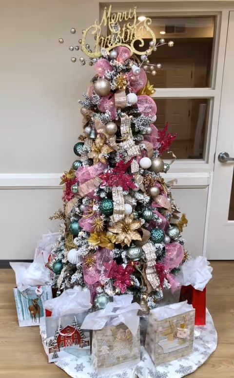 A decorated Christmas tree with pink, gold, green, and white ornaments, ribbons, and flowers. The tree topper is a gold 'Merry Christmas' sign. Several wrapped gift bags with tissue paper are placed around the base of the tree. The tree is set indoors against a beige wall with a window and a door in the background.
