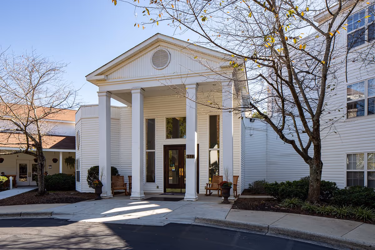 White senior living building entrance with tall columns, a covered portico, benches, and surrounding trees.