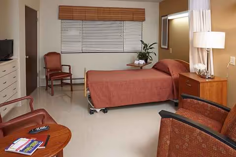 Senior care bedroom with a twin bed, nightstand, dresser, chairs, and window blinds.