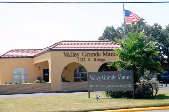 Front exterior of Valley Grande Manor nursing and rehabilitation building with entrance, sign, landscaping, and an American flag.