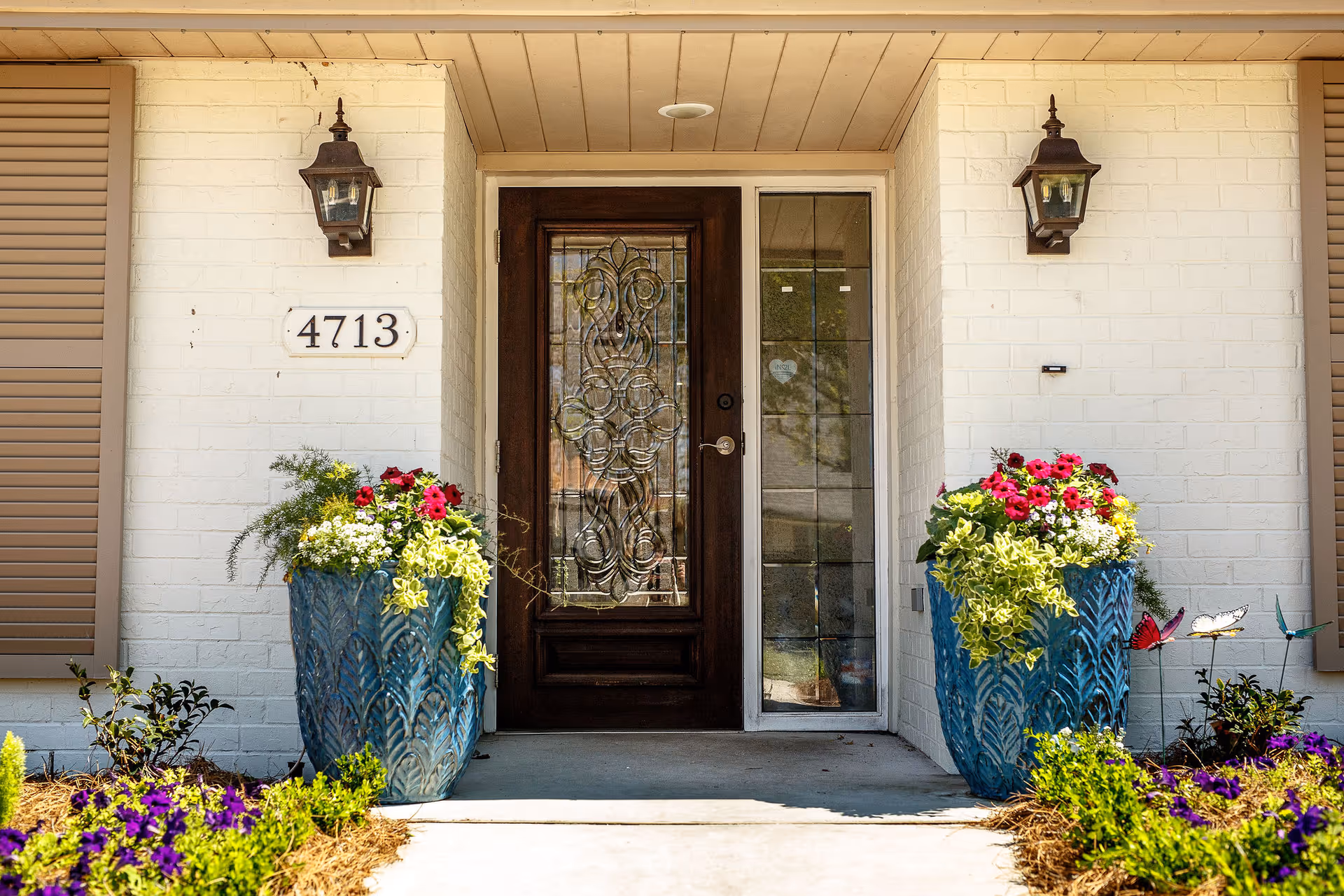 Front entrance of a building with a dark wooden door featuring decorative glass panels. The door is flanked by two large blue planters filled with colorful flowers and greenery. The building has white brick walls, two wall-mounted lantern-style lights, and the number 4713 displayed on a white plaque to the left of the door. There are flower beds with purple flowers on either side of the walkway leading to the door.