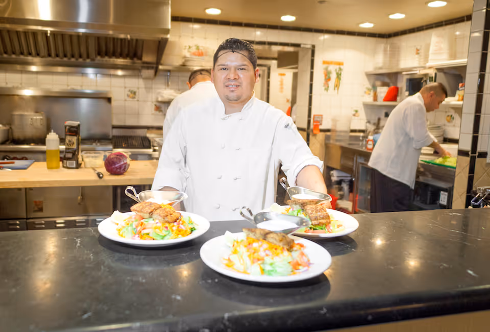 A chef in a white uniform stands behind a counter in a commercial kitchen, presenting three plates of food with grilled meat and colorful salads. Another chef is working in the background preparing ingredients.