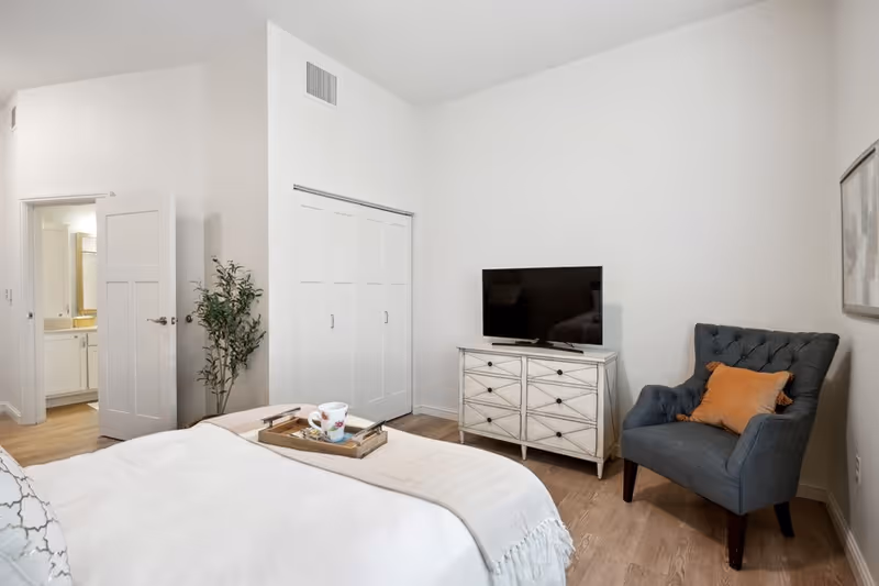 A bright bedroom with a white bedspread and a tray holding a cup and a book on the bed. There is a gray upholstered armchair with an orange pillow to the right, a white dresser with a flat-screen TV on top, and a closet with white sliding doors. A doorway leads to a bathroom with a sink and mirror visible. The walls are white and the floor is wood.