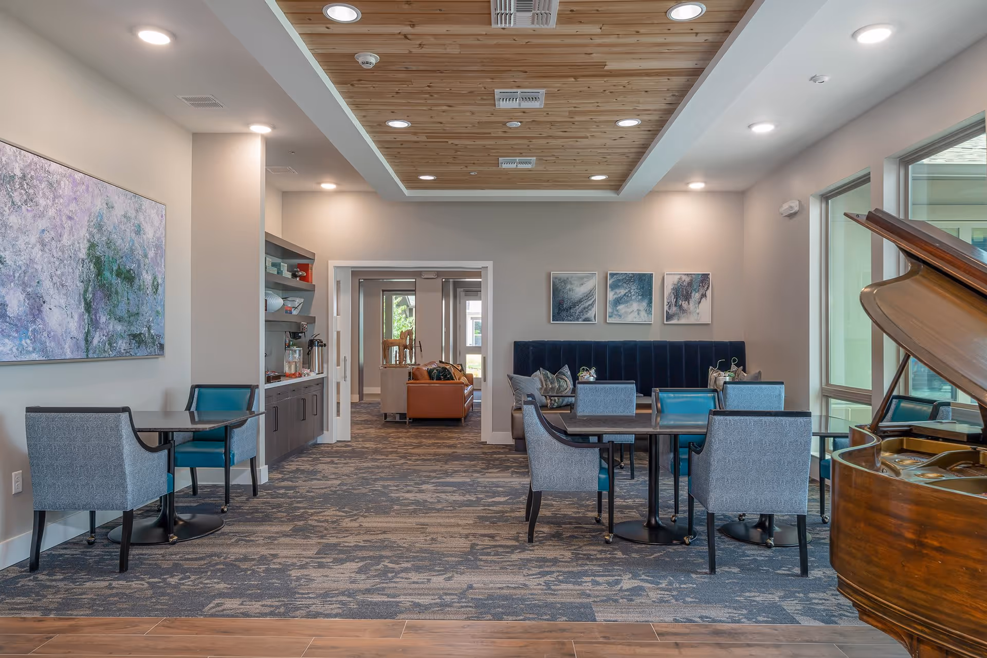 A modern and well-lit common area in a senior living facility featuring multiple tables with blue and gray chairs, a wooden grand piano on the right, abstract artwork on the walls, and a wooden ceiling panel with recessed lighting. In the background, there is a seating area with a brown leather couch and additional chairs.