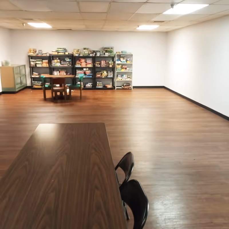 A spacious room with wooden flooring and white walls, featuring a long wooden table with two black chairs in the foreground. In the background, there are several shelves filled with board games and a small table with three chairs.