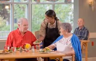 A caregiver serving food to two elderly people seated at a dining table in a well-lit room with large windows. Another elderly man is seated in the background.