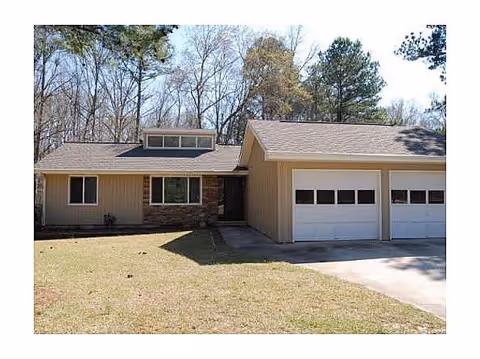 Single-story house with beige siding and stone accents, featuring a two-car garage and a small front lawn with trees in the background under a clear sky.