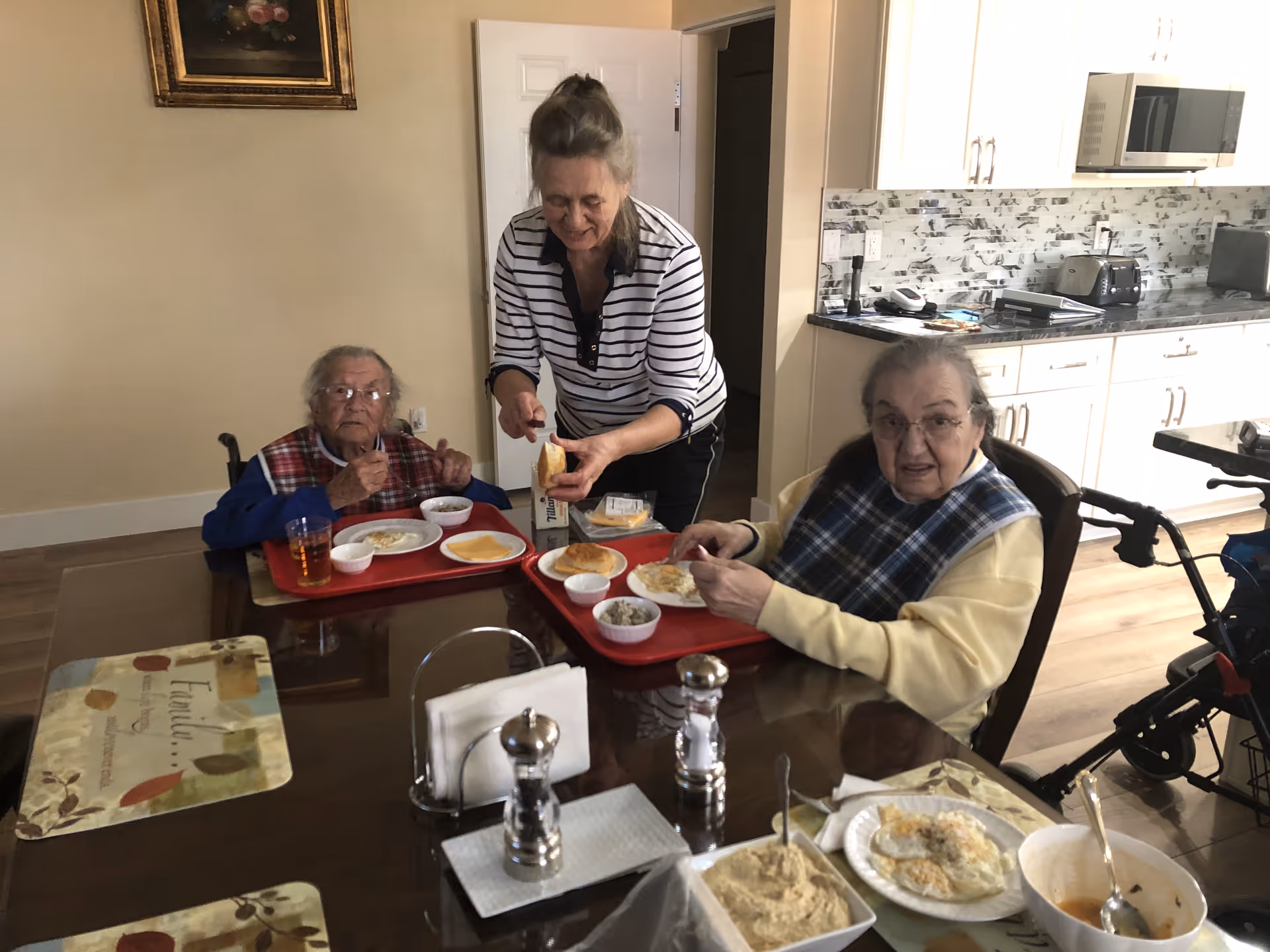 Two elderly women sitting at a dining table with red trays of food, including eggs and bread, while a caregiver stands between them, assisting with the meal. The setting is a kitchen area with white cabinets and a microwave in the background.