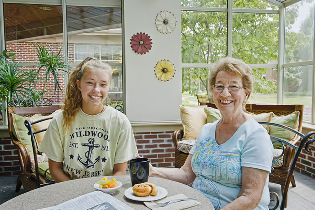 An elderly woman and a young woman sitting at a round table in a sunroom or enclosed patio area. The elderly woman is wearing glasses and a light blue shirt with a floral design, smiling at the camera. The young woman is wearing a beige Wildwood New Jersey t-shirt and also smiling. On the table are a plate with a donut, a bowl of fruit, a black mug, and a newspaper. The background shows large windows with a view of greenery outside and decorative wall hangings.