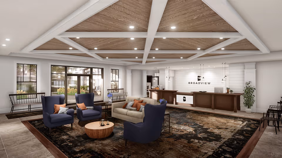 A spacious and modern senior living facility lobby with a wooden coffered ceiling, recessed lighting, and large windows letting in natural light. The seating area includes two blue armchairs, a beige sofa with orange and patterned pillows, and a round wooden coffee table on a large patterned rug. The reception desk is in the background with the sign 'BROADVIEW' on the wall behind it. There are benches along the walls and some potted plants adding greenery to the space.