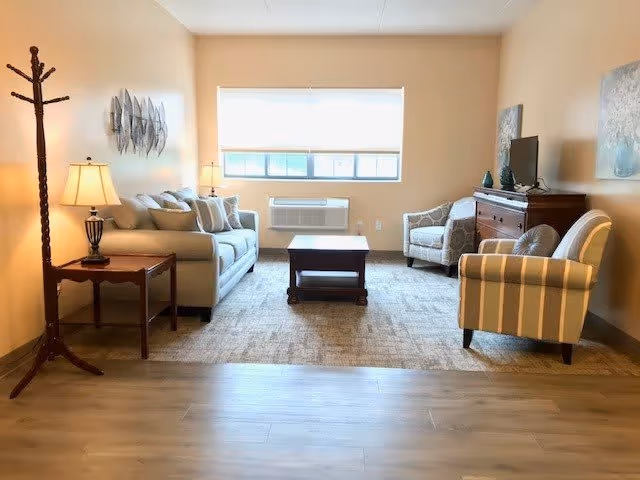 A bright living room with a beige sofa, two armchairs, a wooden coffee table, and a wooden side table with a lamp. There is a coat rack on the left side and a flat-screen TV on a wooden dresser on the right. A large window with a white blind lets in natural light, and the walls are decorated with artwork.
