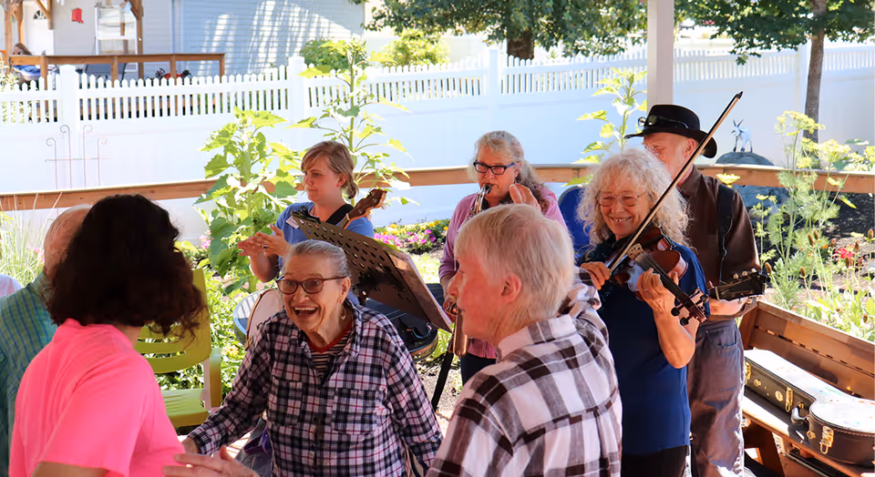 A group of elderly people enjoying a lively outdoor gathering under a covered patio. Some are dancing and smiling, while others are playing musical instruments including a violin, clarinet, and guitar. The setting is bright and sunny with greenery and a white picket fence in the background.