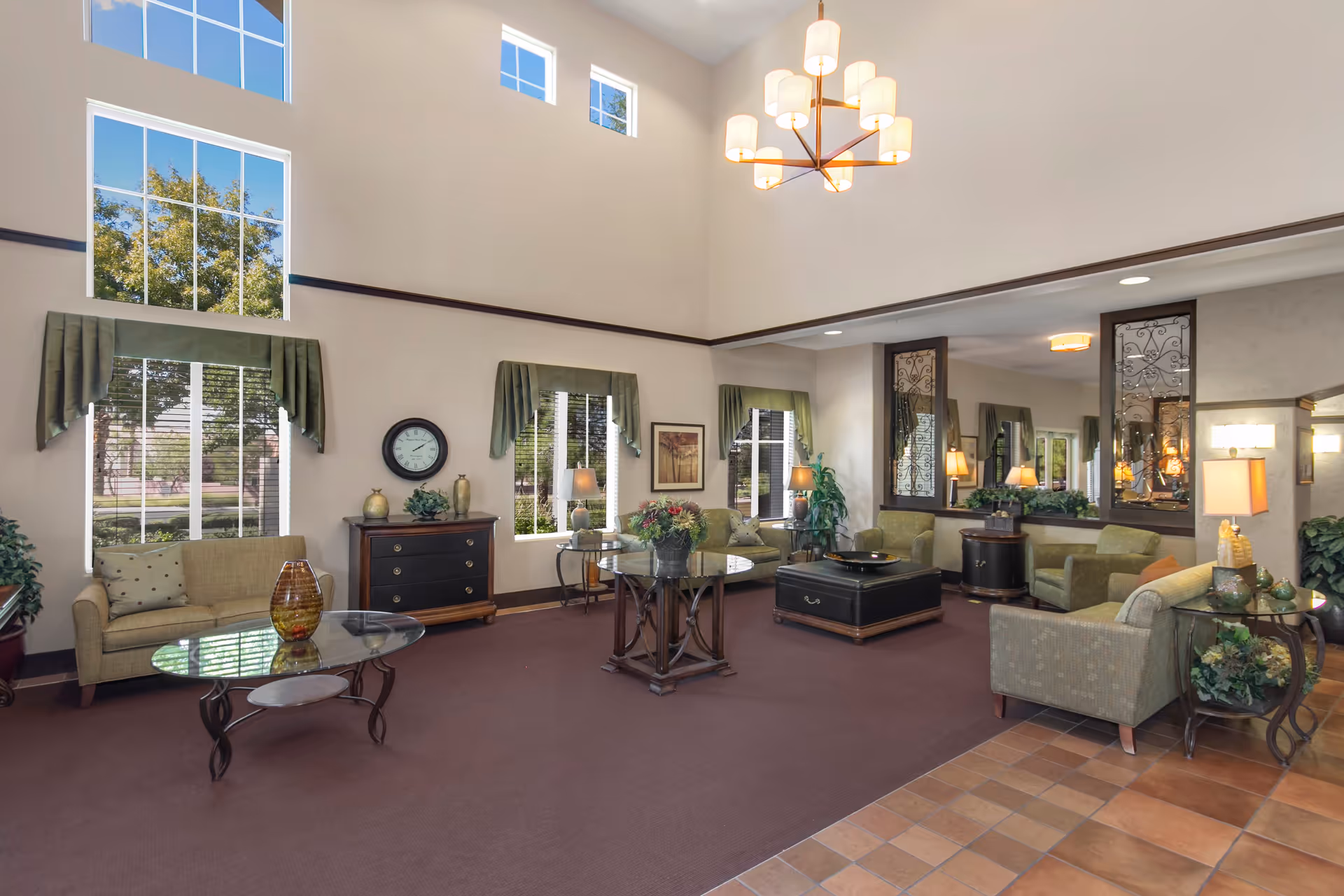 A spacious, well-lit living room area with high ceilings and large windows letting in natural light. The room is furnished with multiple green upholstered chairs and sofas arranged around glass and wooden tables. There are decorative plants, lamps, a clock on a wooden cabinet, and framed artwork on the walls. The floor is carpeted with a section of tiled flooring near the entrance, and a modern chandelier hangs from the ceiling.