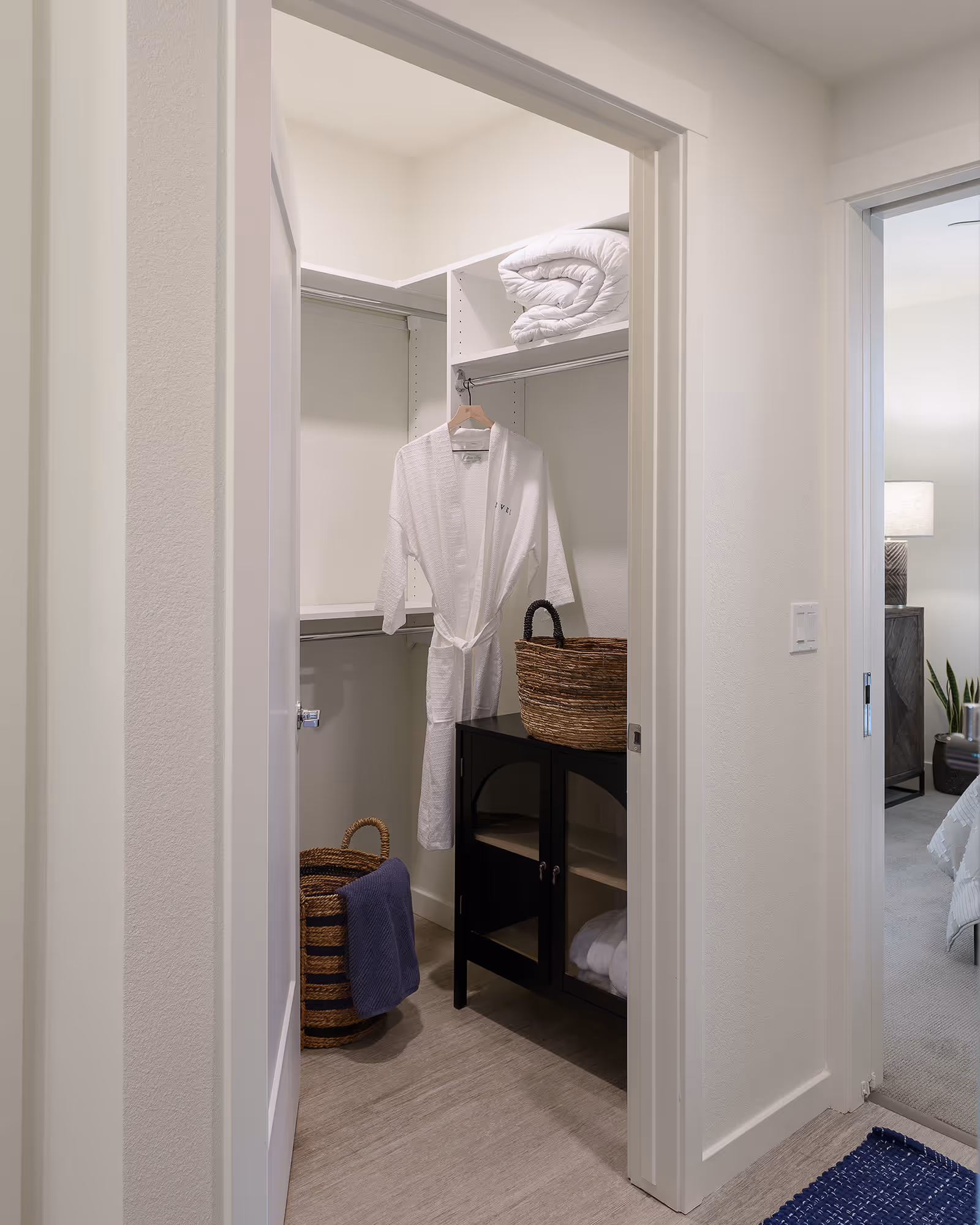 View into a walk-in closet with a white robe hanging on a rod, a rolled-up white comforter on a shelf, a woven basket on a black cabinet, and another woven basket with a blue towel on the floor. Adjacent to the closet is a glimpse of a bedroom with a bed, nightstand, lamp, and plant.