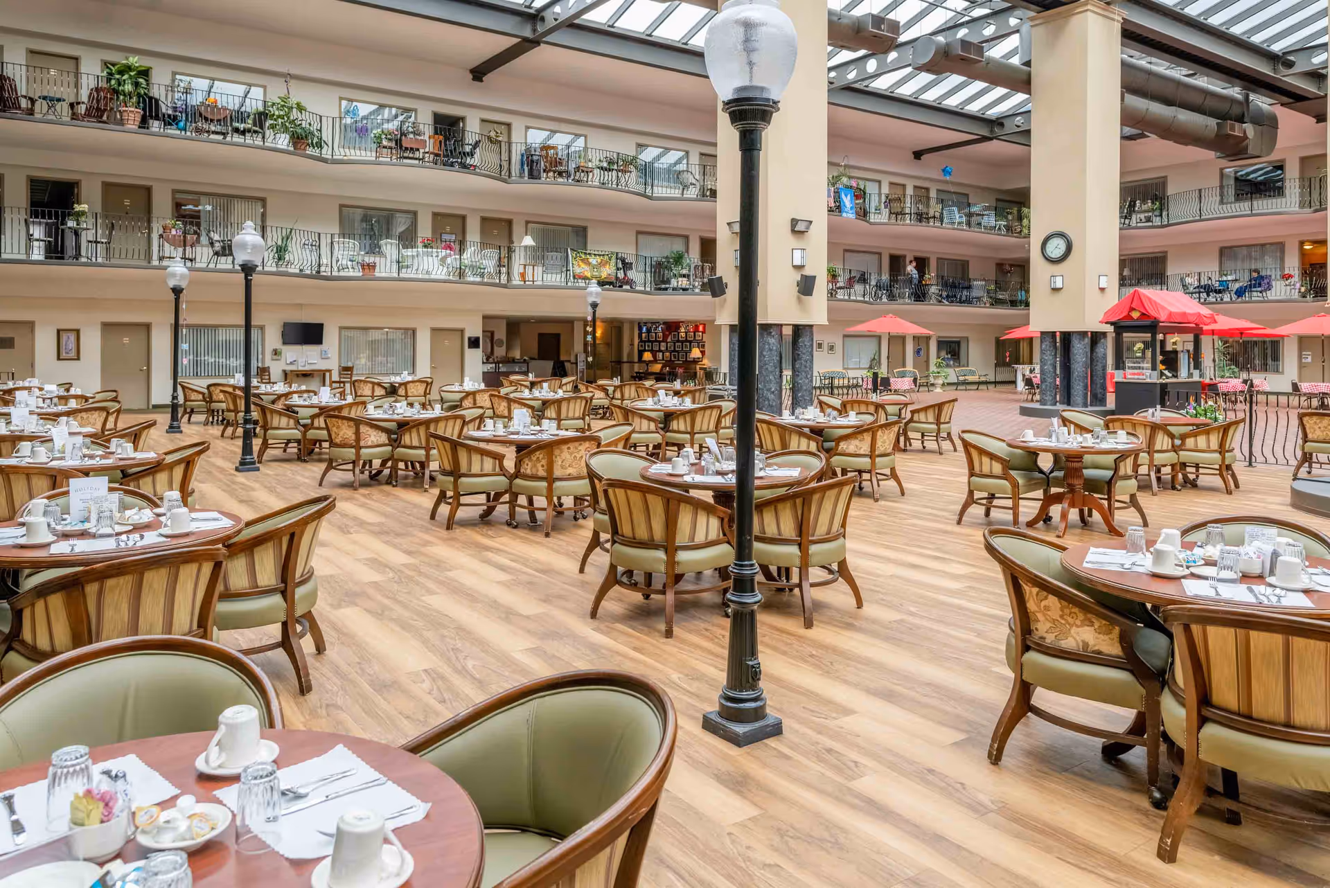 Spacious indoor dining atrium with many round tables and chairs set for meals beneath a glass skylight and surrounded by balcony walkways.