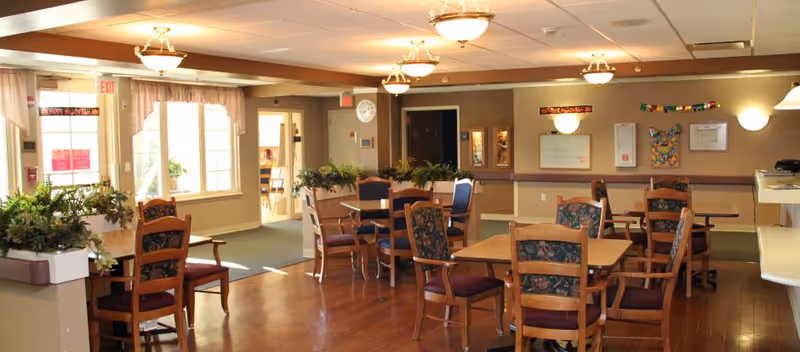 A well-lit common area in Highland Healthcare Center with wooden floors, several wooden tables and chairs with floral upholstery, potted plants on ledges, and wall decorations including a clock and bulletin boards. Large windows allow natural light to enter the room.