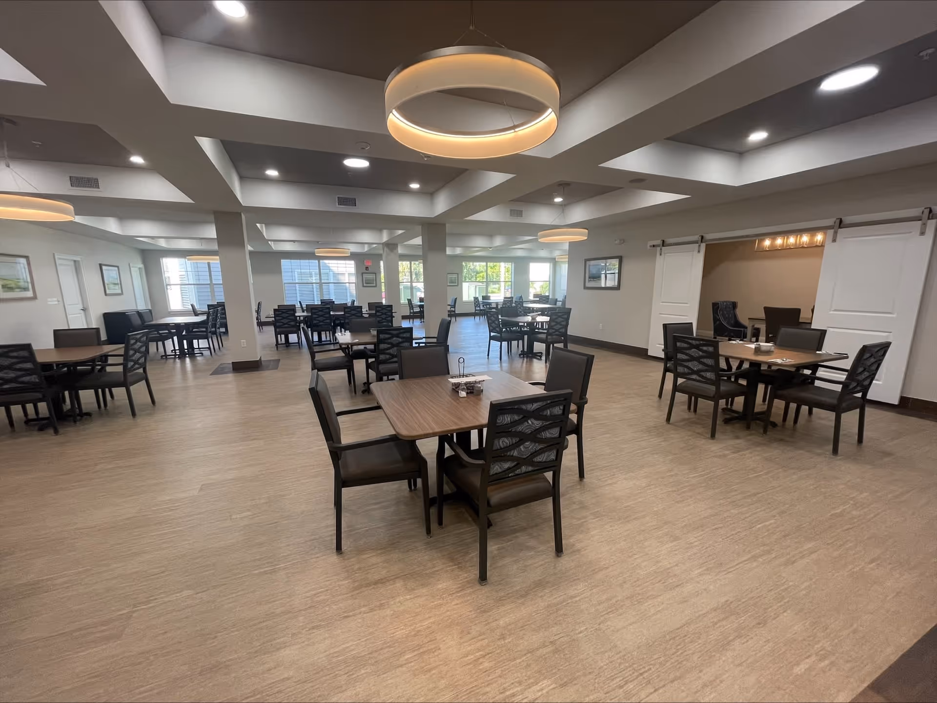 Spacious dining room with multiple square wooden tables and black chairs arranged neatly. The room has large windows allowing natural light, modern circular ceiling lights, and light-colored flooring. There are white sliding barn doors on one side leading to another room.