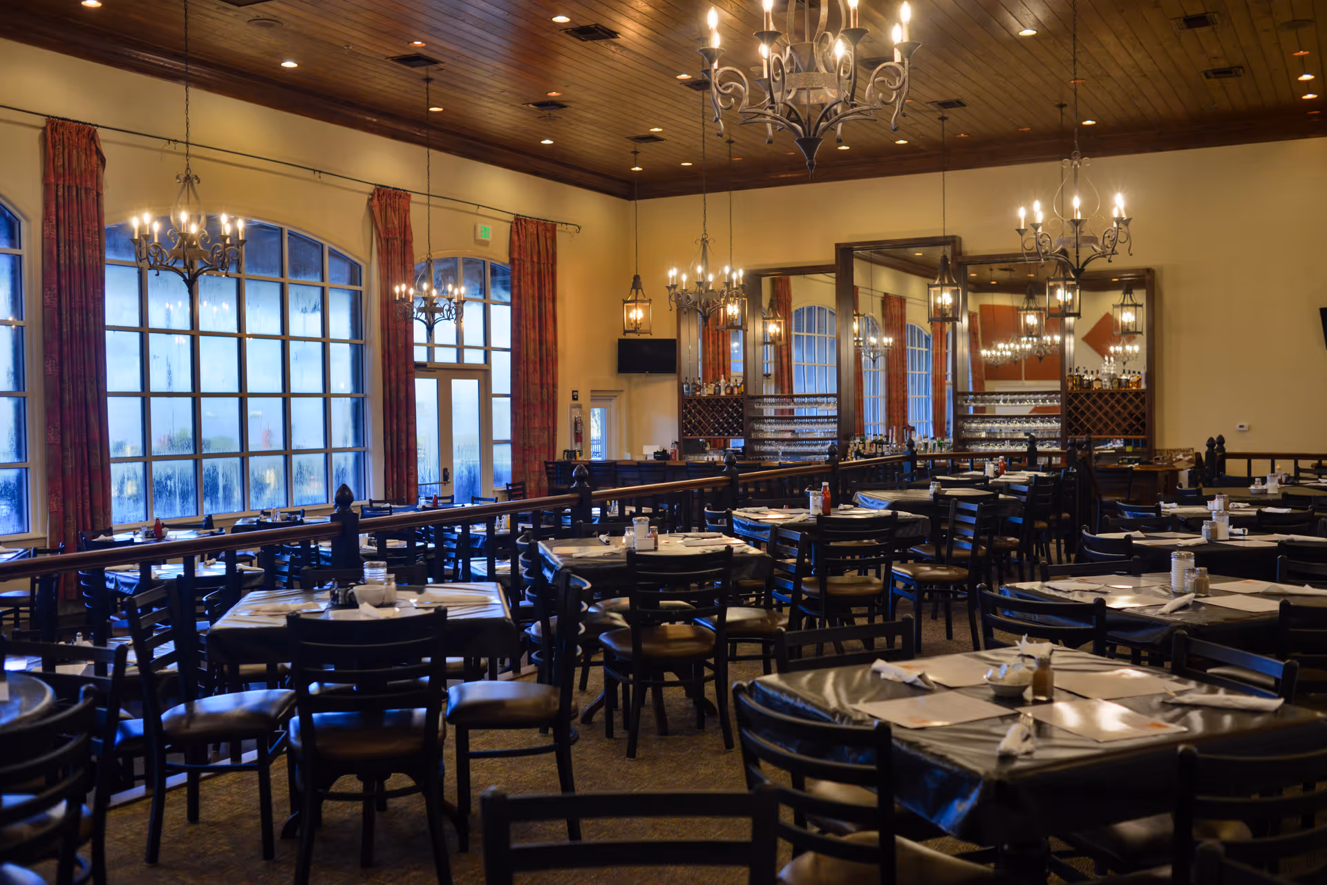 Spacious, warmly lit dining room with multiple set tables, chandeliers, large windows and a bar area.