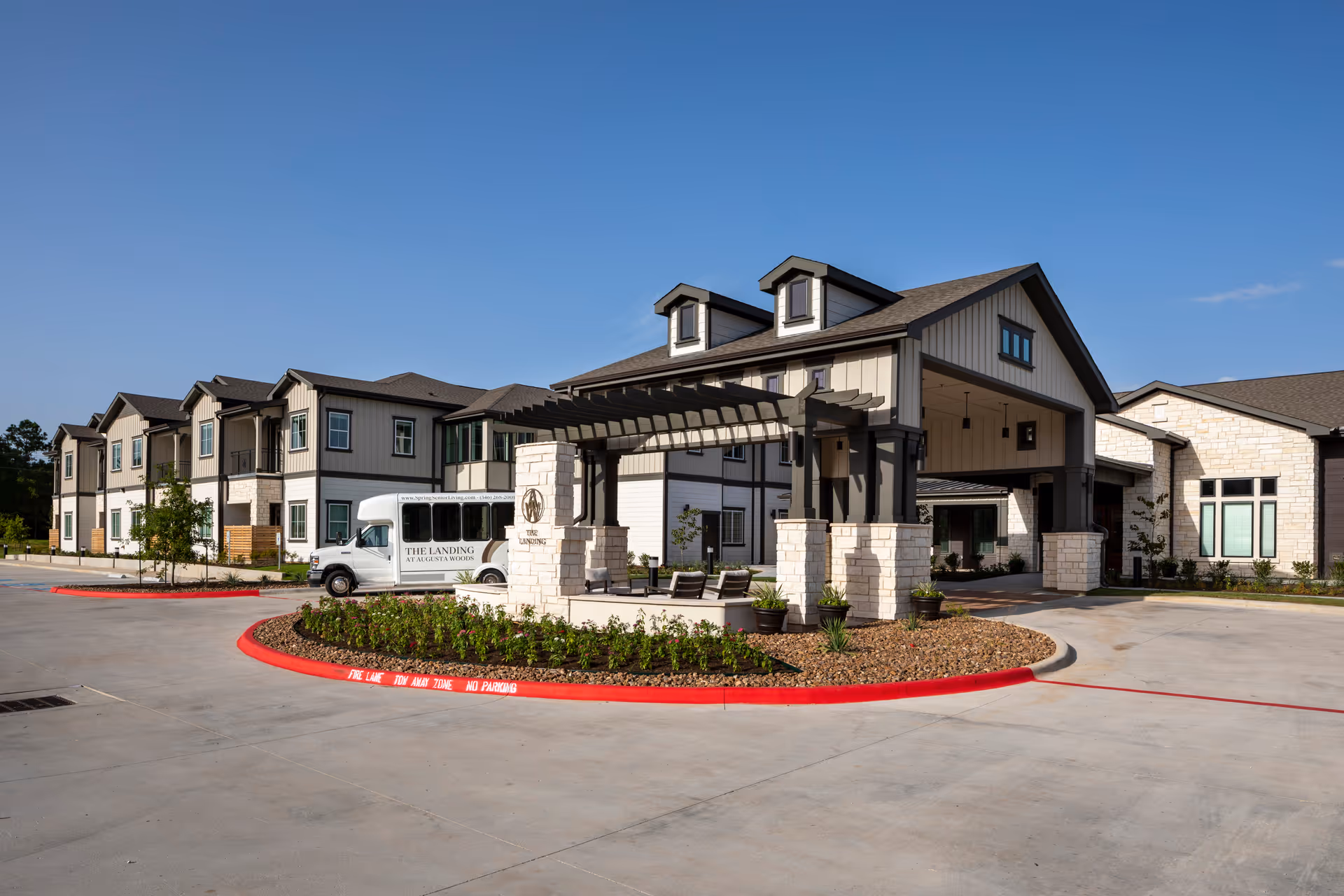 Front entrance of a senior living facility showing a covered porte-cochere, landscaped roundabout, and a shuttle van parked nearby.