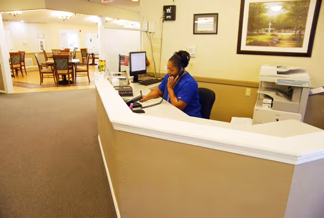 A woman in a blue uniform is sitting at a reception desk, talking on the phone and writing on a notepad. Behind her is a printer and a framed picture on the wall. In the background, there is a dining area with tables and chairs.