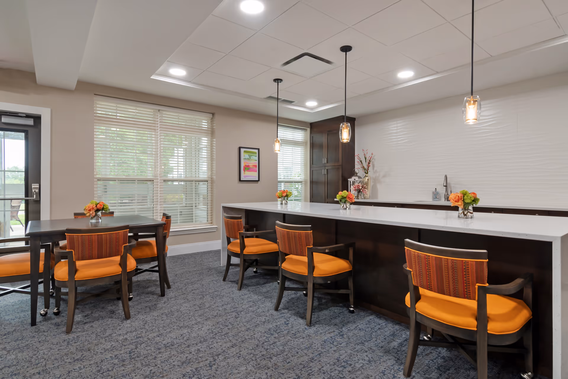 A bright and modern dining area with a long white countertop bar and several wooden chairs with orange cushions. There are pendant lights hanging above the bar, a small table with matching chairs to the left, and large windows with blinds letting in natural light. The room has a carpeted floor and neutral-colored walls with a decorative piece and flowers on the counter.