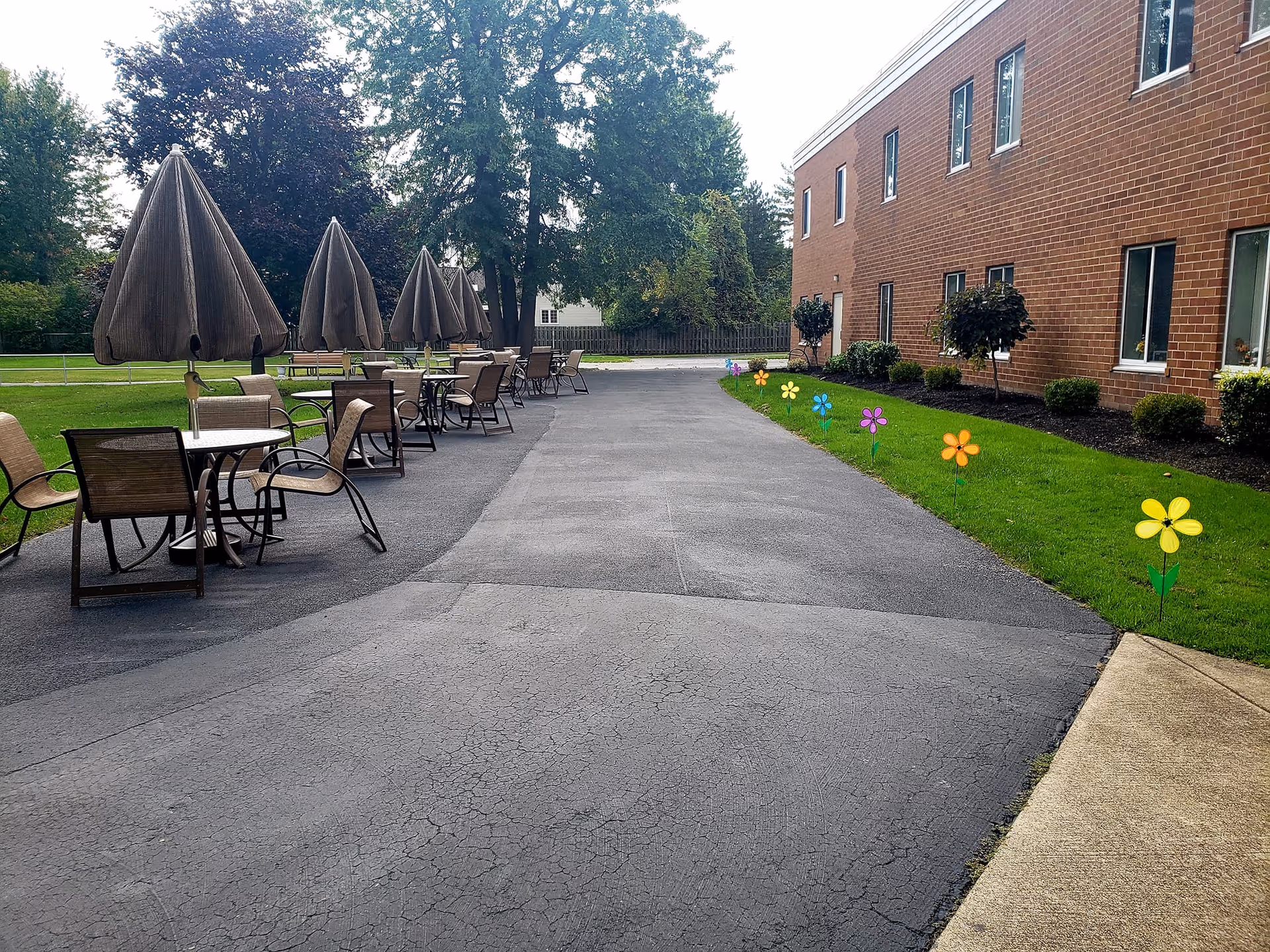 Outdoor patio area at Heathwood Assisted Living and Memory Care with several tables and chairs, each table shaded by a closed umbrella. The patio is adjacent to a brick building with windows, and a row of colorful flower-shaped decorations lines the grass along the building. Trees and greenery are visible in the background.