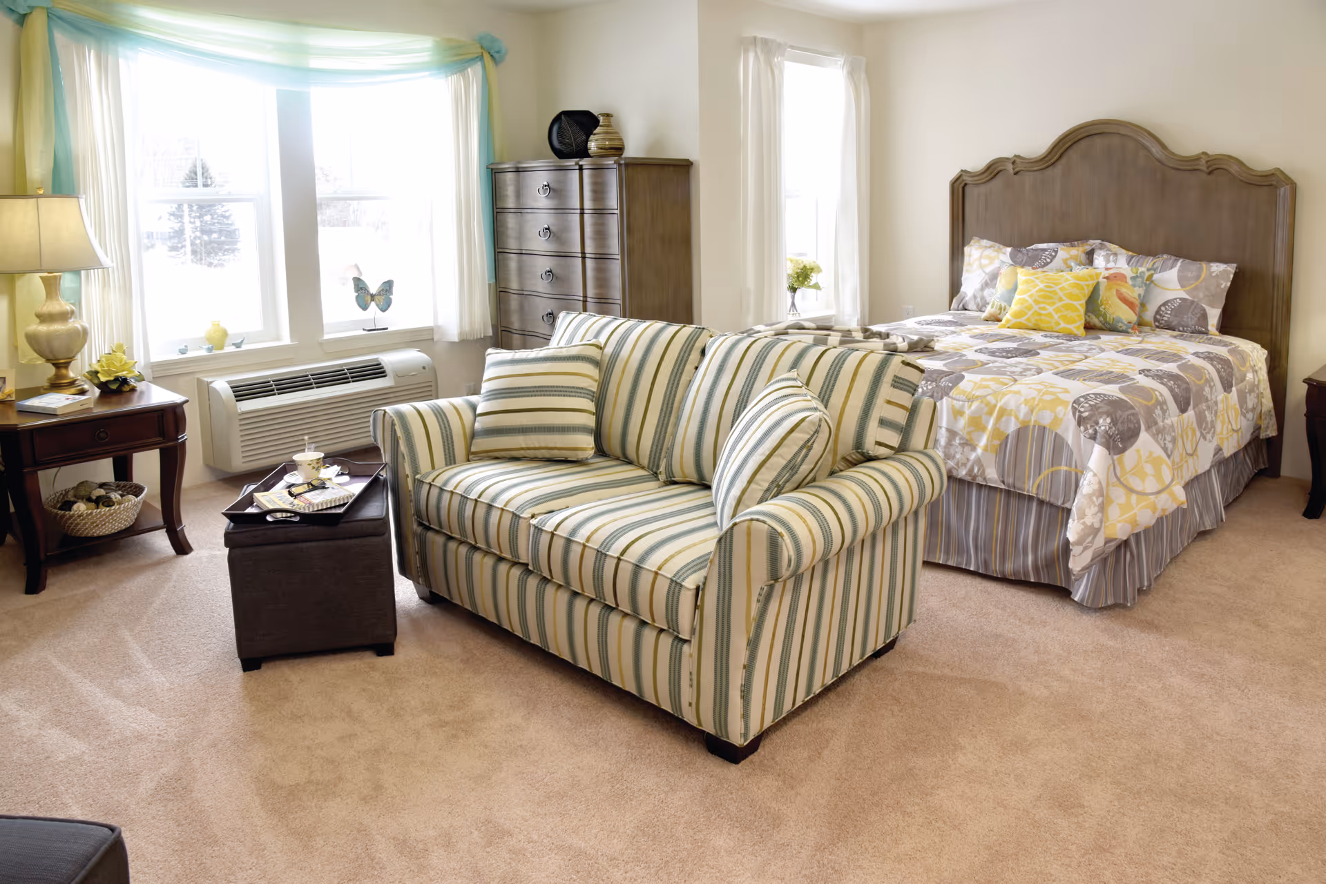 Studio apartment bedroom with a striped loveseat, patterned bed, dresser, and large windows letting in light.