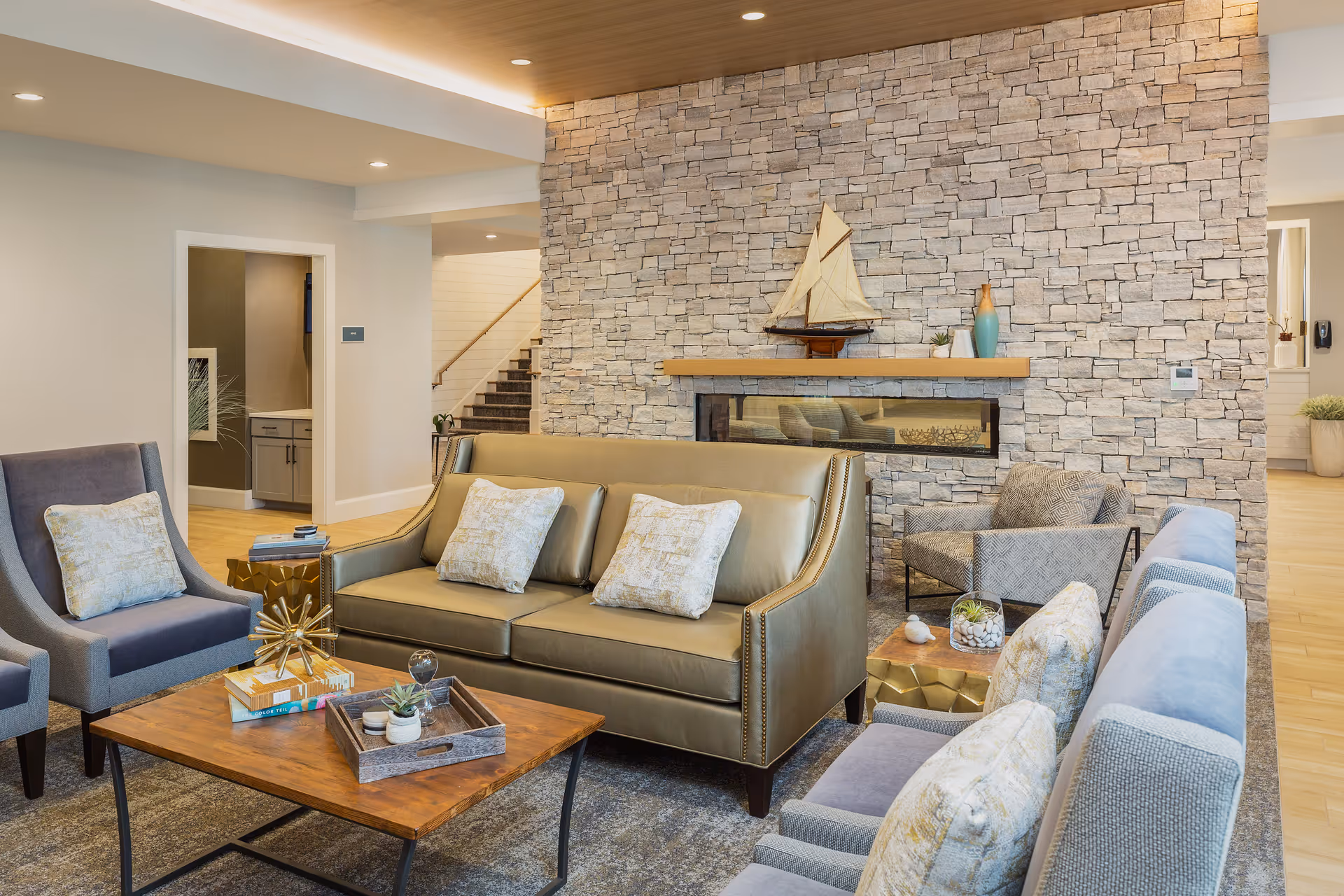 A cozy living room area with a stone accent wall featuring a modern horizontal fireplace. The room has a beige leather sofa with three patterned cushions, two gray armchairs with cushions, and a wooden coffee table with decorative items including books and small plants. A model sailboat and vases are displayed on the wooden mantel above the fireplace. The room has warm lighting and a staircase is visible in the background.
