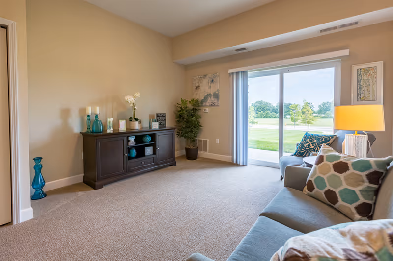 A cozy living room with beige walls and carpet, featuring a dark wooden cabinet decorated with candles, vases, and small framed signs. There is a potted plant in the corner and a large sliding glass door that opens to a green outdoor area. A gray sofa with patterned pillows and a side table with a lamp are visible on the right side.