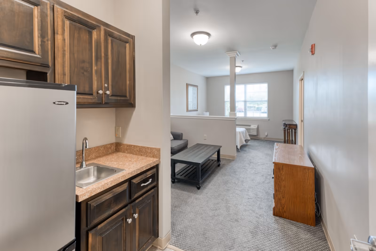 Interior view of a senior living facility unit showing a small kitchenette with a stainless steel mini fridge, sink, and wooden cabinets on the left. The room extends into a living area with a coffee table, sofa, and a bed near a window with blinds. The space is carpeted and has neutral-colored walls with ceiling lights.