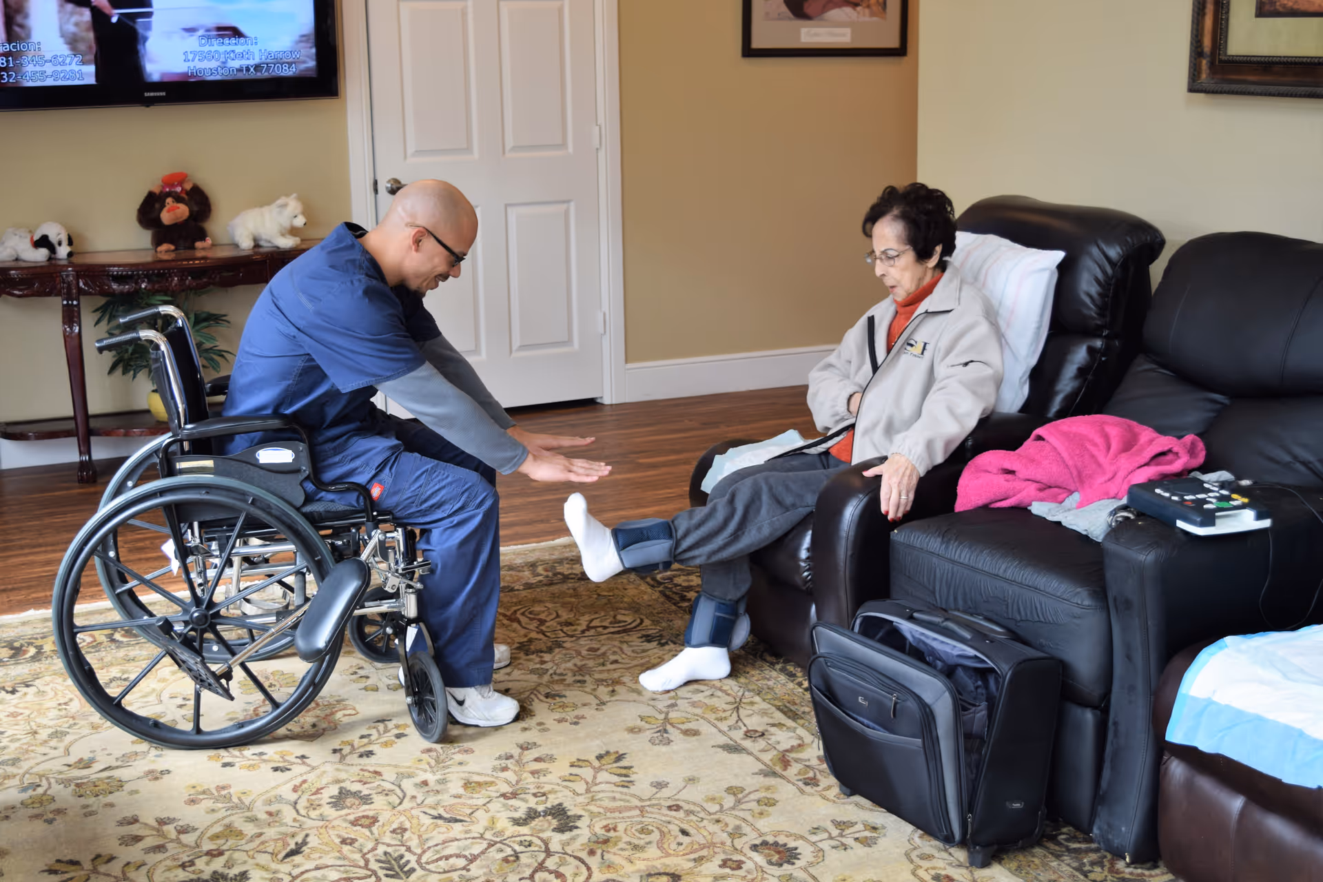 A caregiver in blue scrubs sitting in a wheelchair is assisting an elderly woman seated on a black leather recliner. The woman is wearing a beige jacket and has ankle weights on both legs. They are in a cozy living room with a patterned rug, a TV mounted on the wall, and a table with stuffed animals in the background.