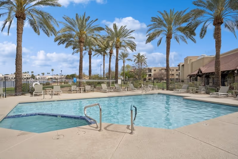 Outdoor swimming pool area with a hot tub adjacent to it, surrounded by palm trees and lounge chairs. The sky is blue with some clouds, and there are buildings and a fence in the background.