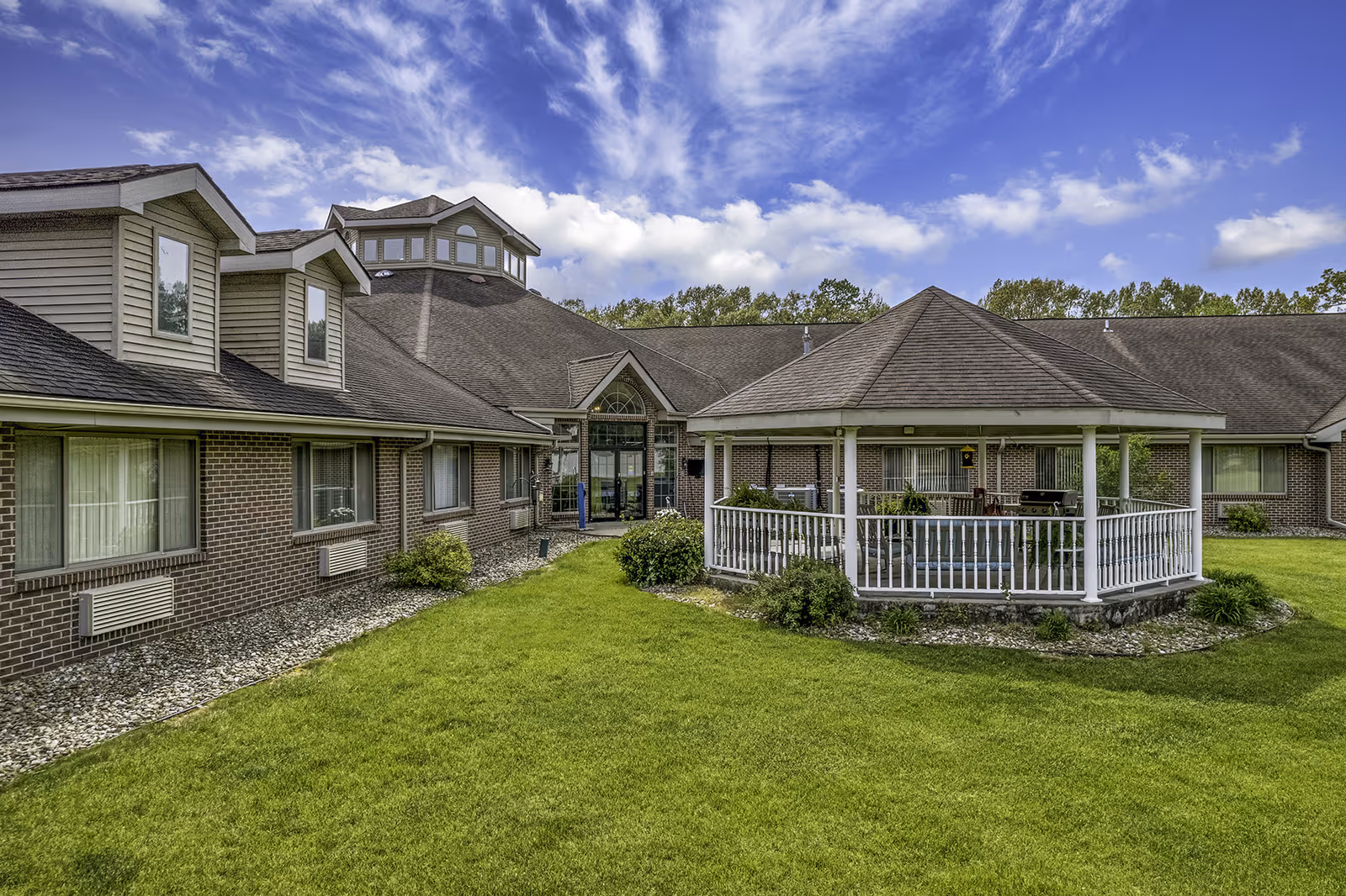 Exterior view of Brentwood at Elkhart Assisted Living facility showing a brick building with multiple windows, a central entrance with glass doors, and a white gazebo surrounded by green grass and shrubs under a partly cloudy blue sky.