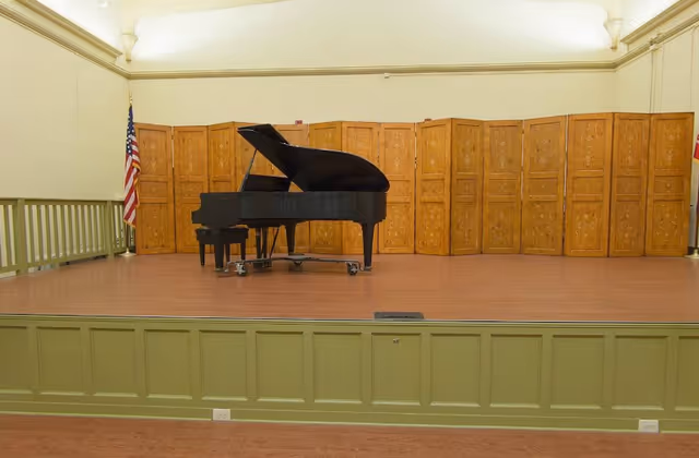 A black grand piano with a matching bench is positioned on a wooden stage with a green paneled front. Behind the piano is a decorative wooden folding screen, and an American flag is visible to the left side of the stage. The room has cream-colored walls and a high ceiling with soft lighting.