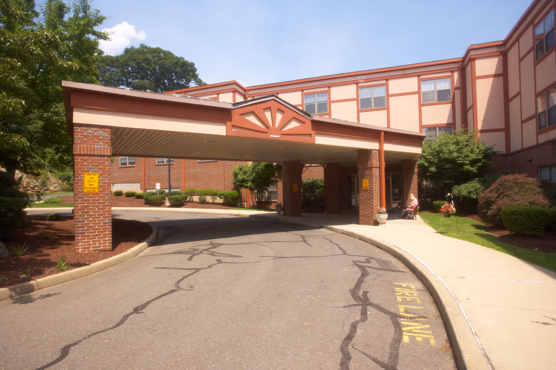 Entrance of a brick building with a covered drop-off area supported by brick columns. There is a curved driveway with 'FIRE LANE' painted on the asphalt and a sidewalk with greenery and bushes alongside. A person is sitting on a bench near the entrance under the covered area.