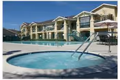 Outdoor view of a senior living facility named Abbey Road Villa featuring a large swimming pool and a circular hot tub in the foreground, with a two-story building and patio umbrellas in the background under a clear blue sky.