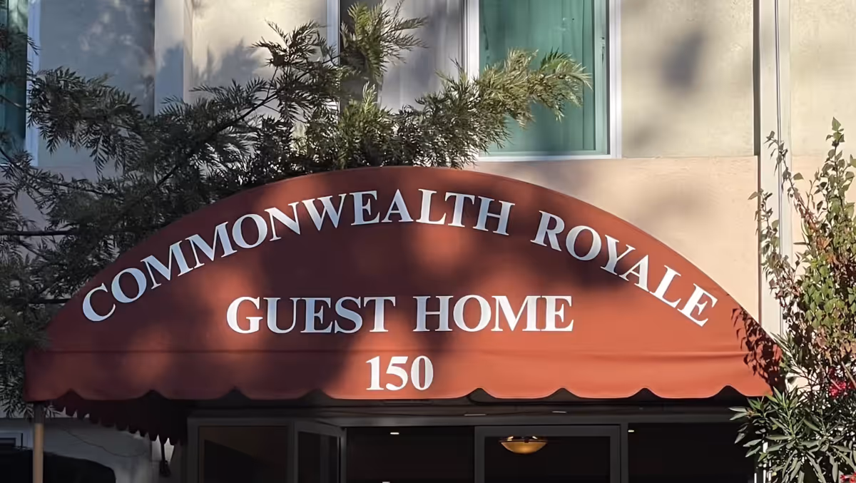 Entrance to Commonwealth Royale Guest Home with a red awning displaying the facility name and number 150, surrounded by greenery and part of the building facade visible.