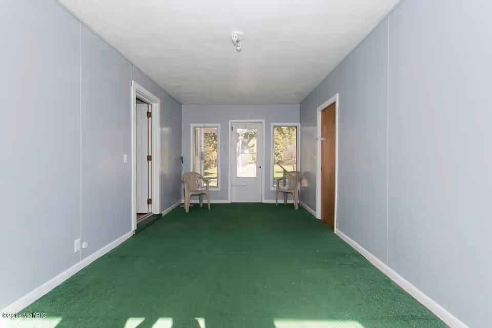 A narrow interior room with light gray walls and green carpet. There are two plastic chairs placed near a door with two windows on either side, allowing a view of the outdoors. Two doorways are visible on the left and right walls.